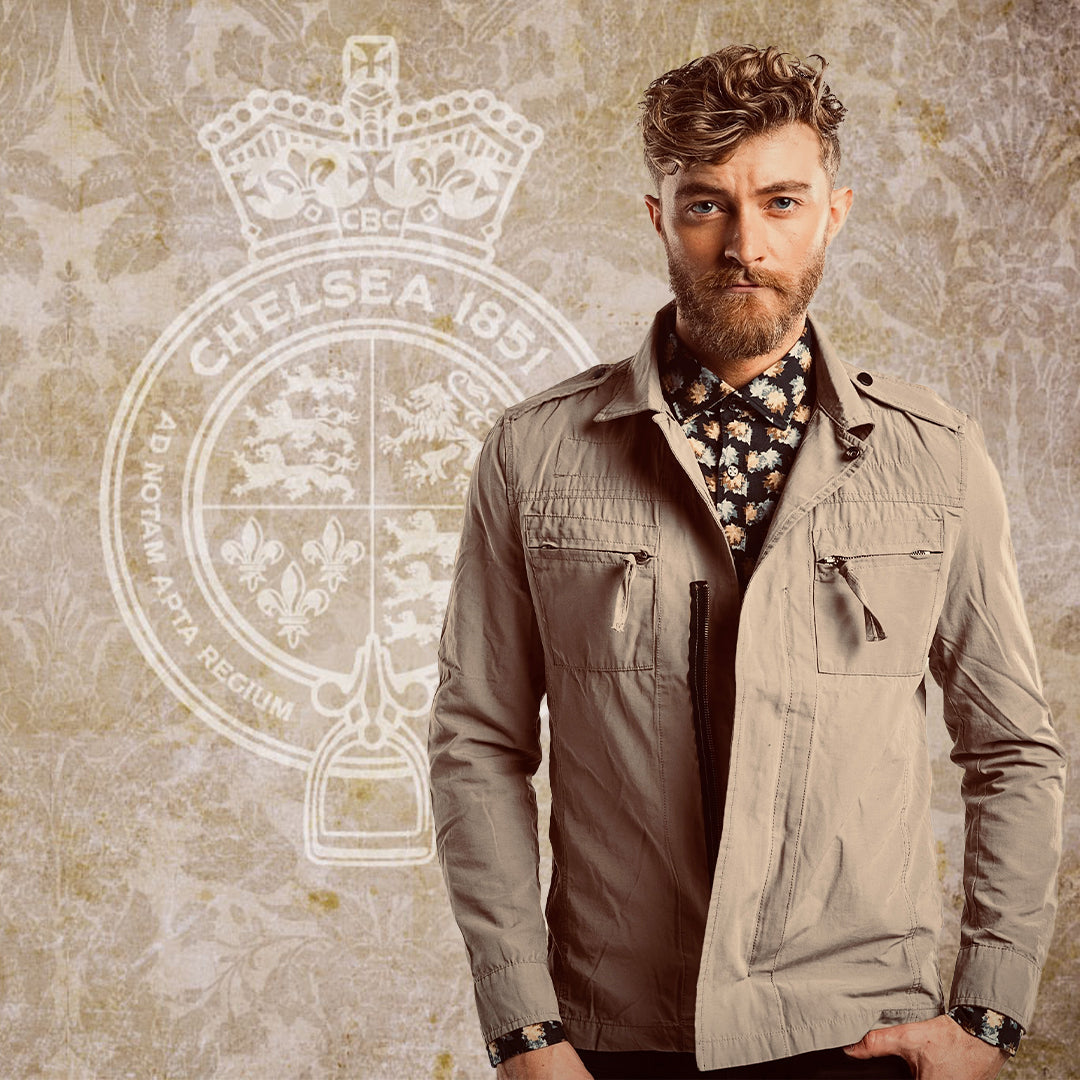 A bearded man with curly hair wearing a light khaki Chelsea Harbour jacket layered over a floral button-up shirt. He stands confidently in front of a vintage-style Chelsea Boot Co. crest.